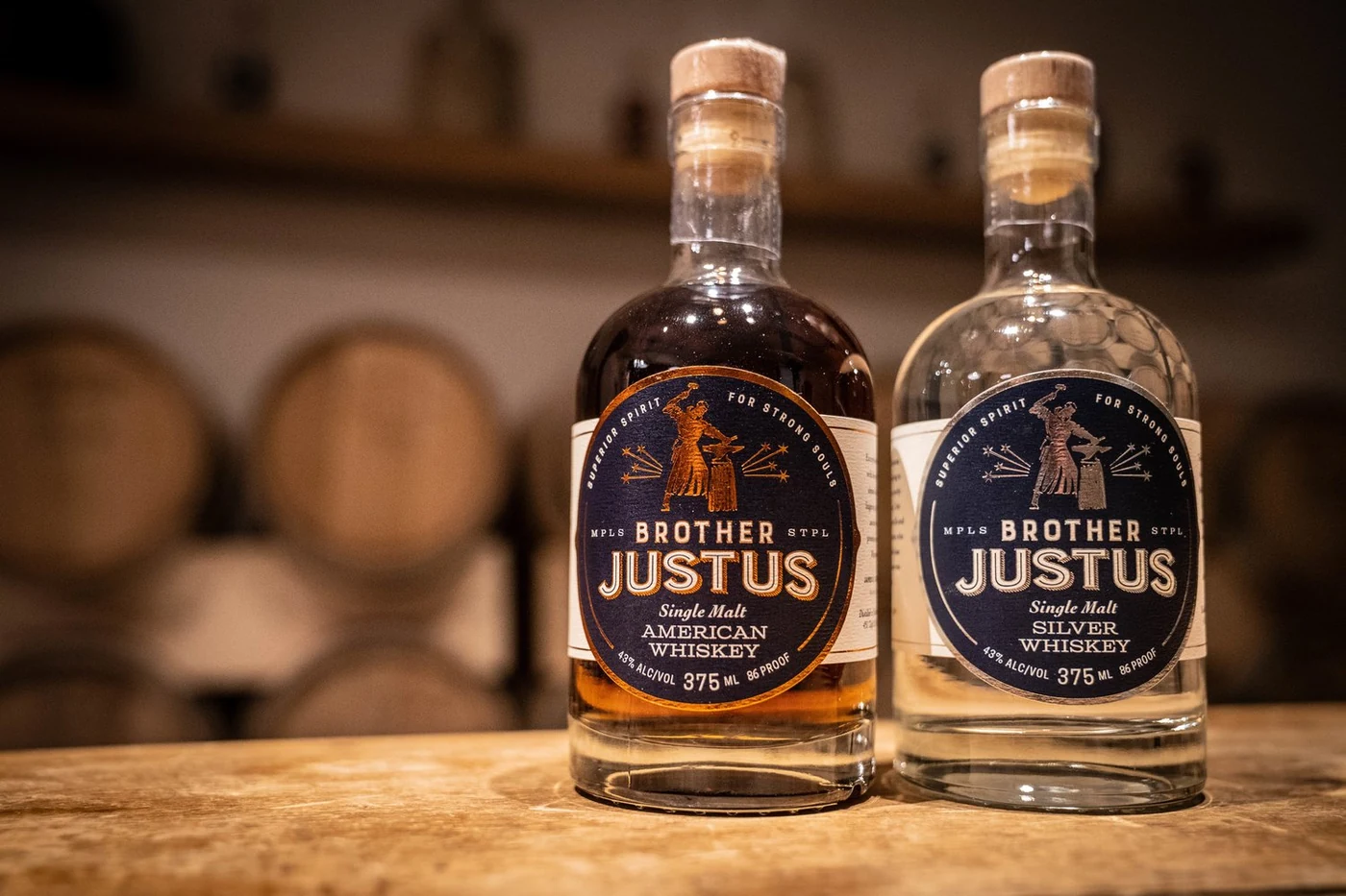 Brother Justus American Whiskey and Silver Whiskey bottles with monk-blacksmith label design, barrels blurred in the background